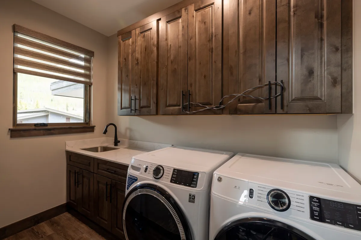 A bright, tidy laundry area featuring a modern washer and dryer, ample wooden cabinetry for storage, and a convenient sink for hand-washing adventure gear. The organization and efficiency of the space are evident, making laundry tasks straightforward and accessible.