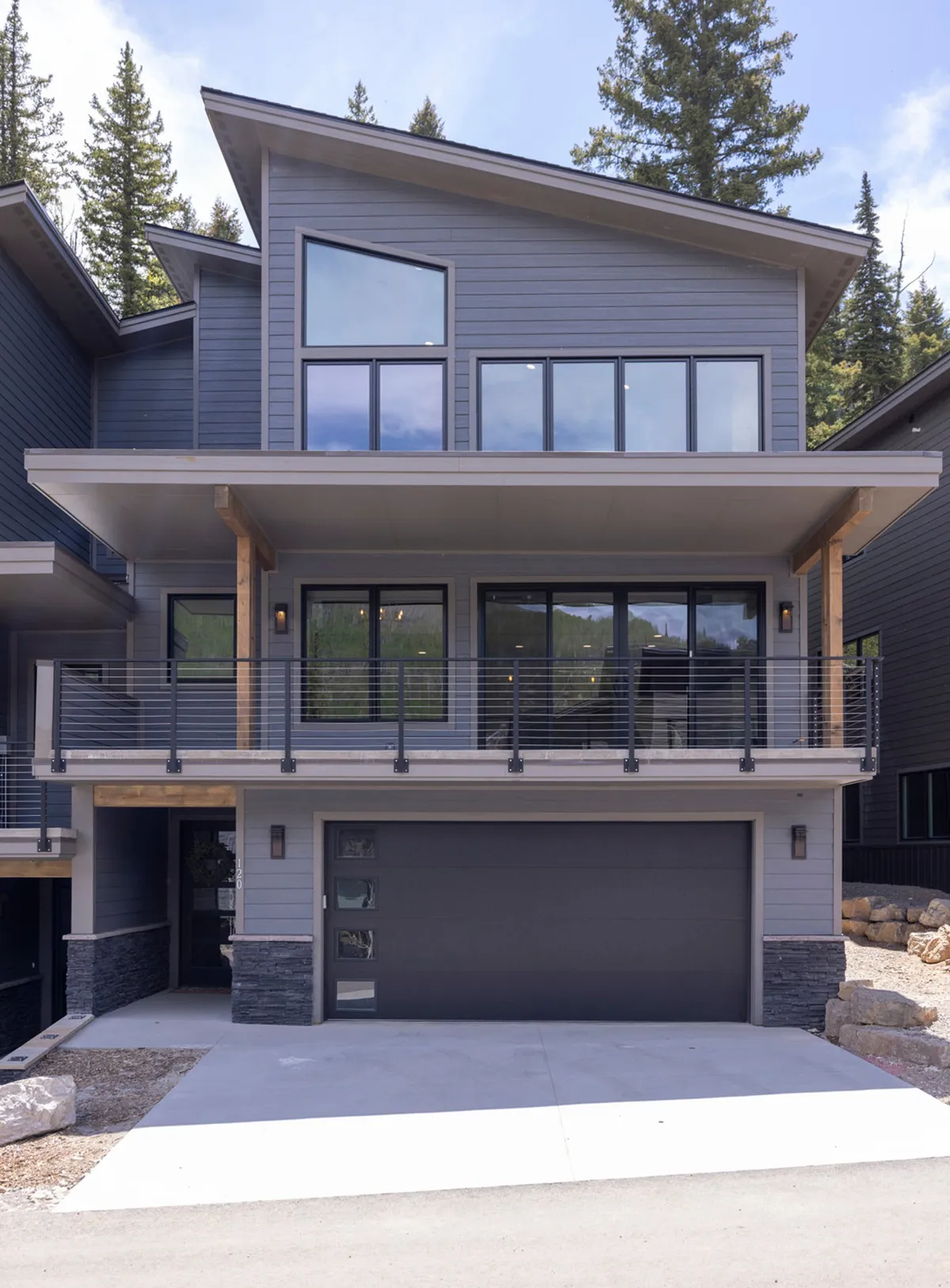 A striking exterior shot of the home featuring large windows, a spacious balcony, and modern stone accents. The balcony is visible as an inviting space to relax or gather, set against a scenic backdrop, ideal for family or group activities.