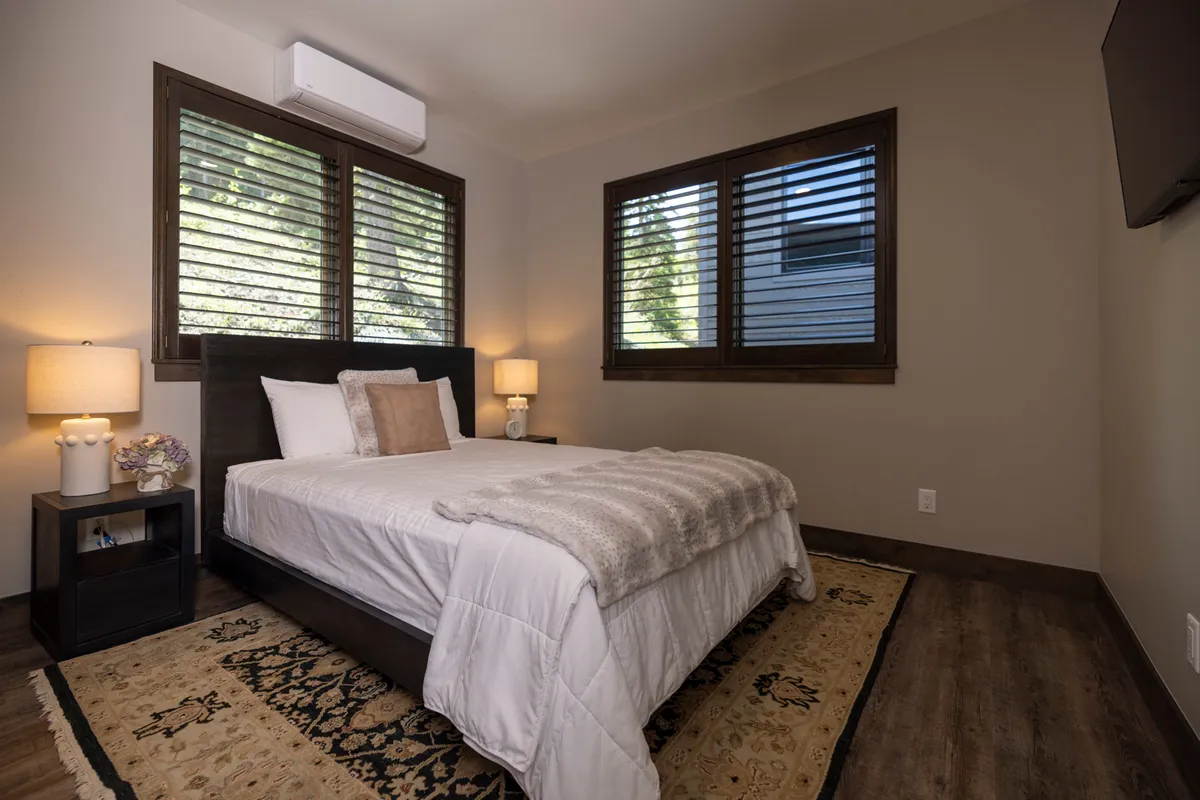 A bright, comfortable bedroom with a large, cozy bed, nightstands, reading lamps, and windows that allow natural light to fill the space. The room's neutral palette and inviting rug contribute to a calm, restful feel.