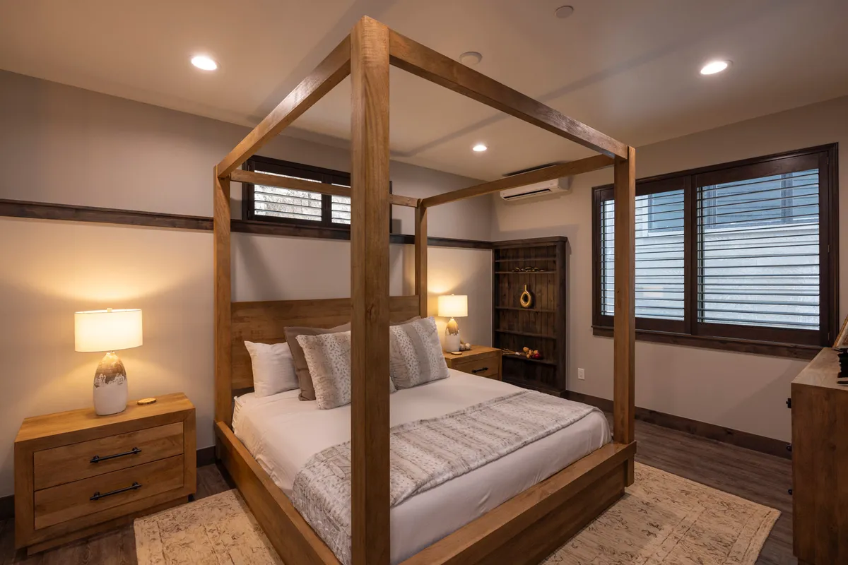 A serene bedroom featuring a four-poster bed flanked by matching nightstands with warm lamps. The room includes a wooden shelving unit and decorative vase, with soft light filtering through the window, creating a peaceful and private retreat.