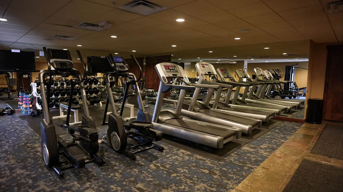 A spacious gym featuring treadmills, ellipticals, dumbbells, and large mirrors, ideal for both individual workouts and group fitness classes. The atmosphere is energetic and communal.