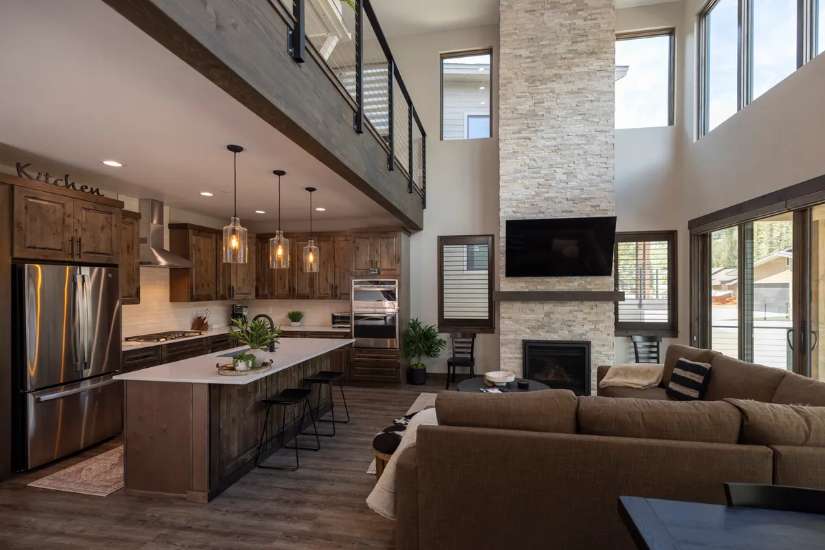 A contemporary kitchen and living area with a large kitchen island, stone fireplace, expansive windows, and an open loft above. The space feels vibrant and inviting—great for socializing, cooking, and entertaining. The kitchen island provides a natural centerpiece, seamlessly connecting the dining and living areas.