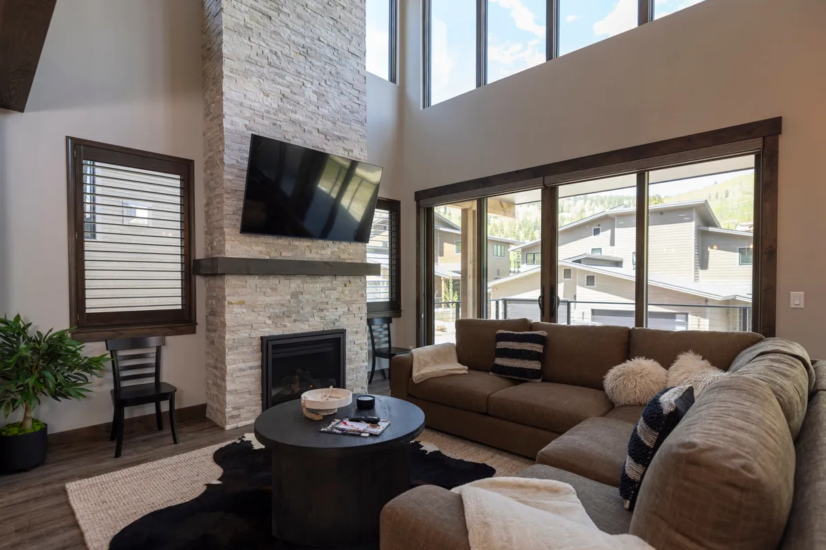 A welcoming open-concept living area featuring a large stone fireplace as the focal point, flanked by a comfortable sectional sofa and a coffee table. Floor-to-ceiling windows bathe the space in natural light and offer glimpses of the surrounding mountains. The atmosphere feels perfect for relaxing with family, movie nights, or hosting friendly gatherings.