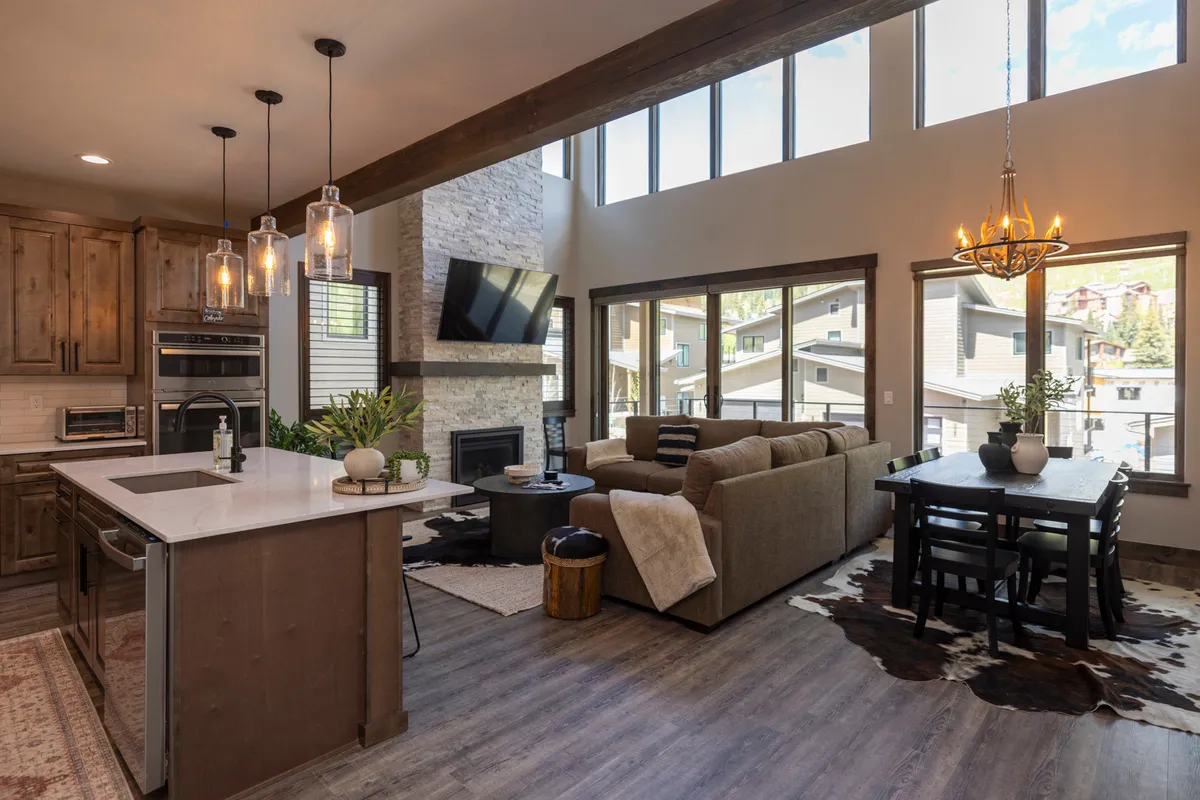 The image showcases a spacious, open-concept living area with a stylish kitchen island, a cozy sofa, an inviting dining table, a modern fireplace, and large windows that fill the space with natural light. The setting is perfect for gathering, entertaining, and enjoying scenic views, reflecting a comfortable and contemporary mountain retreat.