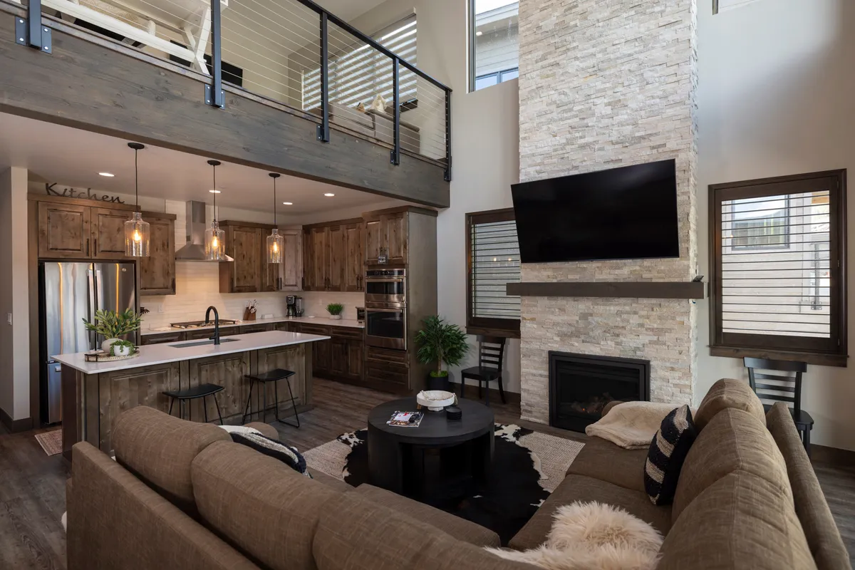 An open plan interior with a stone fireplace, large windows framing scenic views, a modern kitchen with an island, and an upper loft area. The setup is ideal for cooking, relaxing, and gathering, with plenty of natural light and a visual flow between different functional zones.