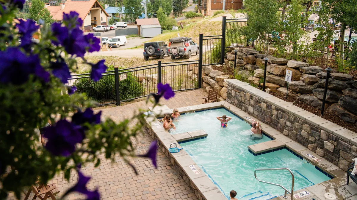 An inviting outdoor scene showcasing a hot tub and swimming pool surrounded by blooming flowers and people enjoying the space. The mood is leisurely and cheerful, with a focus on socializing and enjoying scenic outdoor amenities.